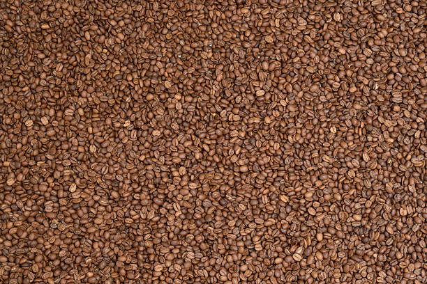 A close-up, top-down view creating a full frame background or texture of freshly roasted coffee beans. Perfect for use as a background or in designs related to coffee, cafes, beverages, food, and energy. Showcases the rich color and texture of whole coffee beans.
