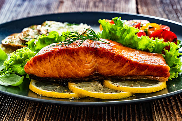 istock Fried salmon steak with fried potatoes, lemon and fresh vegetables served on black plate on wooden background 2214313525