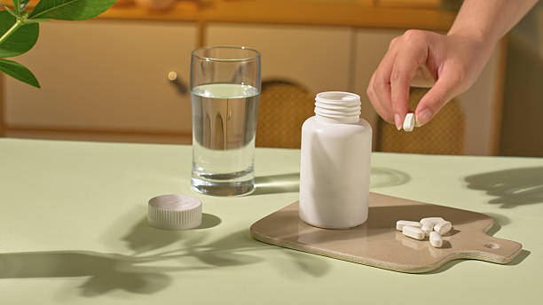 istock A hand is picking up a pill, some capsules placed on the tray, next to it is an open medicine bottle, displayed on a light brown tray. A glass of pure white water and a pot of green plants. 2214319723