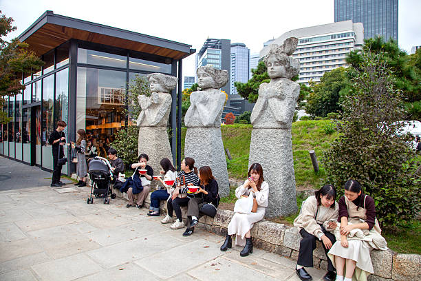 Osaka, Japan - November 17, 2025.\nPeople sitting on a stone wall in Osaka Castle Park, Osaka, Japan with statues behind them.