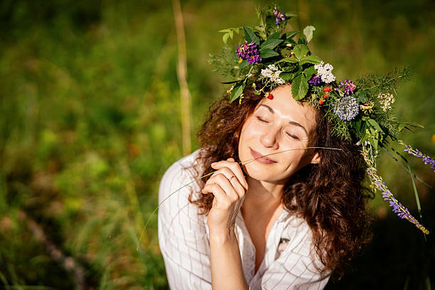 Happy Slavic woman in floral wreath sitting on grass and smiling peacefully in countryside. The adult female model celebrates midsummer with harmony and connection to nature