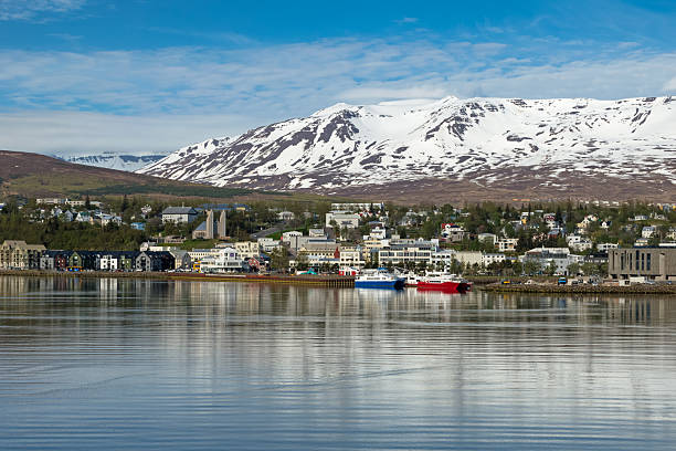 The tranquil waterfront of Akureyri, Iceland, nestles by icy waters beneath towering snow-capped peaks, with a lone boat adding life to the serene winter scene.