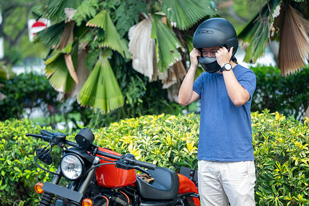 Confident senior man wearing a leather jacket and vintage helmet, fastening the chin strap while sitting on a bench next to his old-school moped. The scene portrays a sense of freedom, independence, and timeless style, blending modern casual fashion with retro motorbike culture. A leather bag and sunglasses complete his adventurous and relaxed look.
