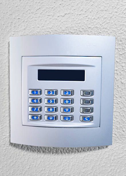 Close-up shot of a modern electronic security keypad mounted on a white textured wall, used for home or office alarm system access control. Blue backlit numeric buttons and display.