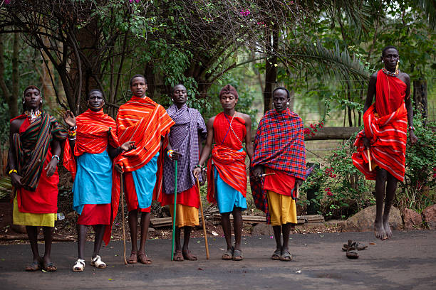 Kenya – April 7, 2012: A group of young Masai men perform a traditional jumping dance outdoors. Dressed in colorful shuka fabrics, they are seen in mid-ritual, with one individual visibly higher in the air than the others. Lush greenery and flowering bushes appear in the background.