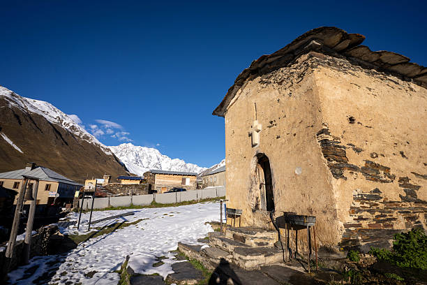 A view captures a weathered, stone church with a simple arched doorway and a stone-slab roof in the remote village of Ushguli, Georgia. Patches of snow cover the ground, and traditional stone houses are visible nearby, all set against a backdrop of majestic, snow-capped mountains under a clear blue sky. This scene highlights the historical and architectural significance of Ushguli, a UNESCO World Heritage site in the Svaneti region.