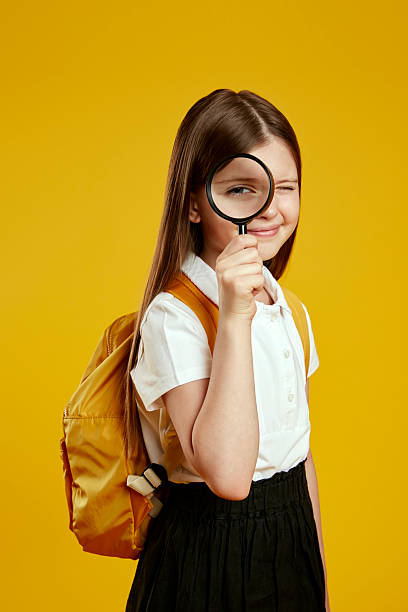 istock Curious young schoolgirl with backpack holding magnifying glass on bright yellow background 2214376717