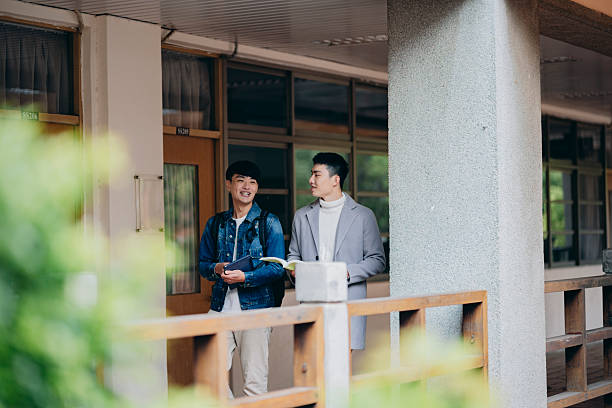A group of Asian university students walk out of a classroom, engaging in lively discussion about coursework. The scene captures the authentic atmosphere of modern campus life, where learning extends beyond lectures and into peer interaction. As academic pressure, tuition costs, and global competition increase, students seek balance between intellectual pursuit and social connection, reflecting how higher education is also a journey of identity, purpose, and future aspirations.