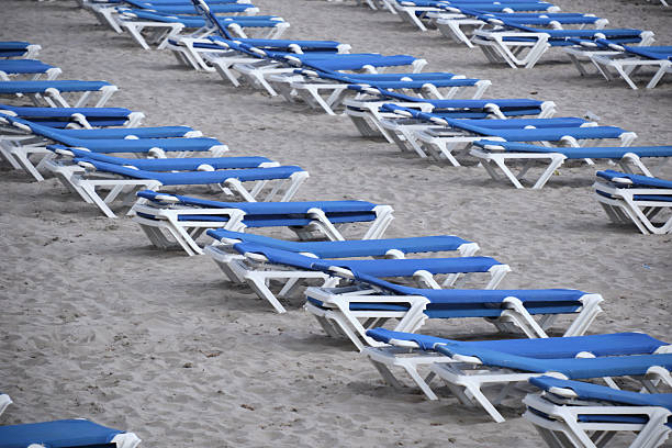 Stacks of white plastic lounge chairs rest on quiet sandy beach, neatly arranged and strapped. Empty shoreline and gentle waves evoke a calm atmosphere awaiting visitors