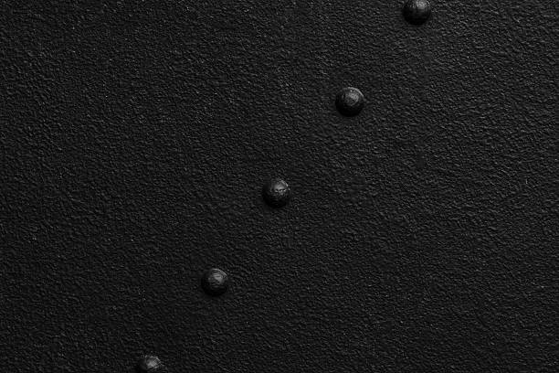 A textured black metal surface features prominent studs, creating an intriguing pattern. The lighting highlights the roughness and depth, enhancing the overall visual appeal.