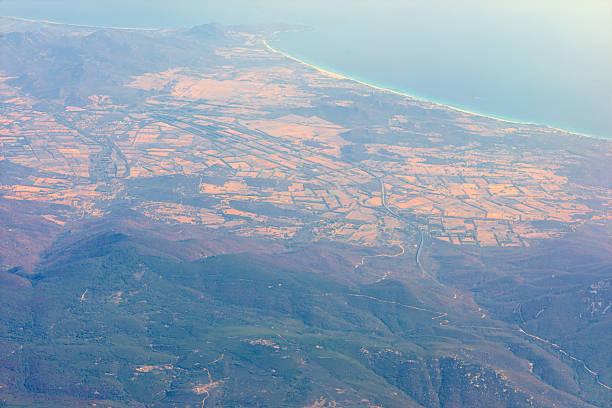 Aerial view of Sardinia with a long, sandy coastline along turquoise sea. Landscape includes a patchwork of fields and rolling hills, with mountains in the foreground under a clear blue sky