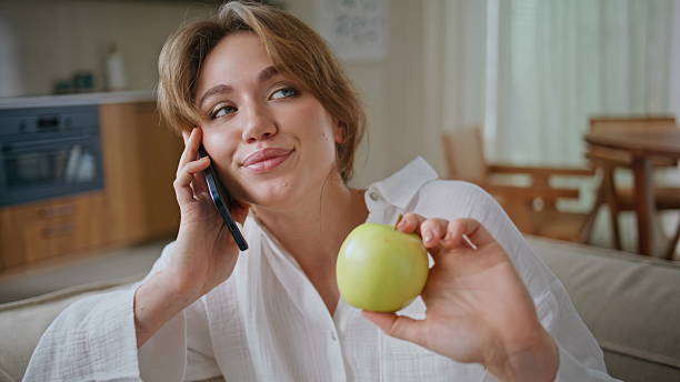 Smiling lady enjoy call on mobile phone relaxing at domestic weekend closeup. Portrait of happy woman talking smartphone at home couch holding apple. Relaxed girl have friendly cellphone conversation