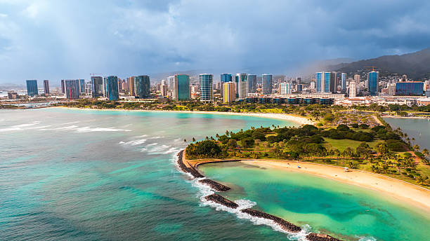 Waves splash by the stone dam protecting the beautiful sandy beach with stunning nature. Aerial view on Waikiki skyline with cloudy backdrop.
