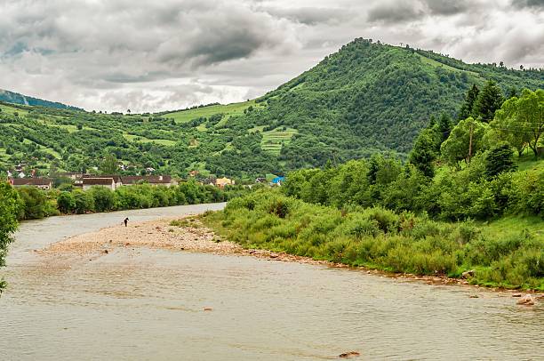 A scenic view of a wide river flowing through a valley with lush green mountains. A small village in the background under a cloudy sky