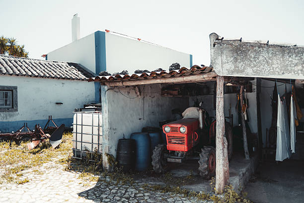 Rustic farmyard scene with a small red vintage tractor parked under a weathered shed, surrounded by traditional whitewashed buildings, cobblestone ground, and farming tools in bright rural sunlight