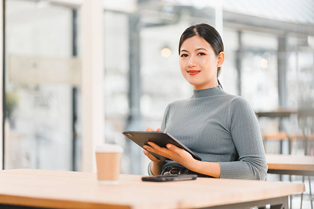 istock Asian woman sitting at table in modern cafe, using tablet and smiling. 2214504070
