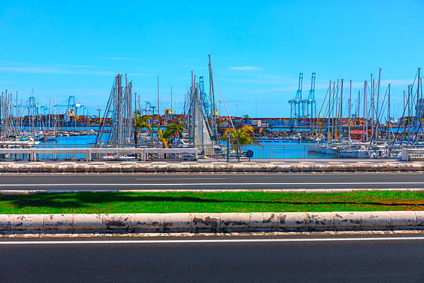 Modern marina in Las Palmas, Gran Canaria, with sailboats docked under a clear blue sky. Industrial cranes and a calm sea backdrop contrast the urban waterfront and palm-lined road
