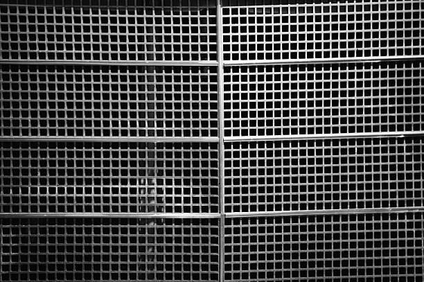 A black and white close-up of a tightly spaced metal grid structure, creating a repetitive geometric pattern. The contrast between light and shadow emphasizes the rigid lines and industrial texture. The image conveys a sense of structure, order, and minimalist abstraction.