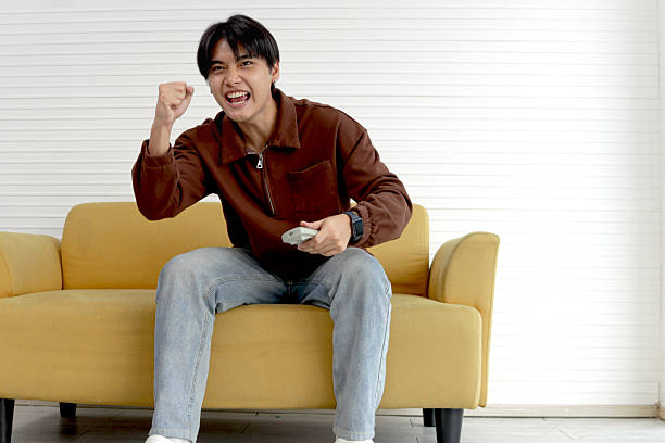 istock Happy young Asian man holding remote control to switch channels while watching movie, sitting on sofa at living room. Smiling teenager raising hand up to cheer  while watching TV show at home. 2214534713
