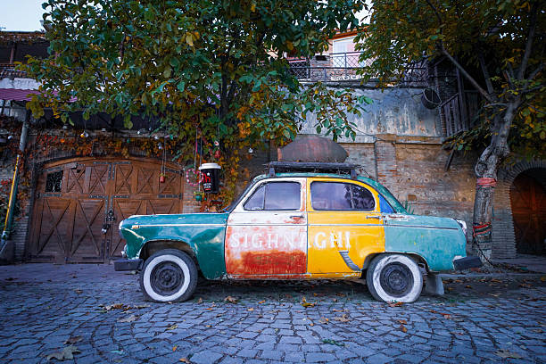 A quirky and colorful scene captures a vintage car parked on a cobblestone street in Sighnaghi, Georgia. The car is painted in a patchwork of teal, white, red, and yellow, with the word \