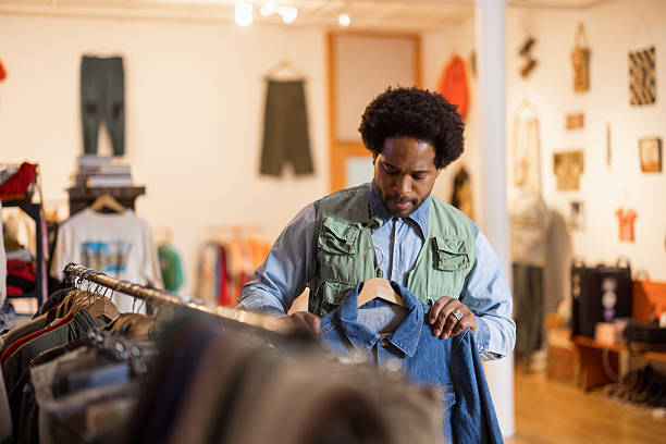 Young adult man browsing through a vintage second-hand shop, searching for the perfect denim shirt among the colorful garments on display
