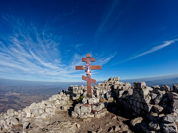 Orthodox cross standing proudly atop a mountain, surrounded by rugged rocks, beneath a vast blue sky, evoking feelings of faith, spirituality, and serene solitude in nature's beauty