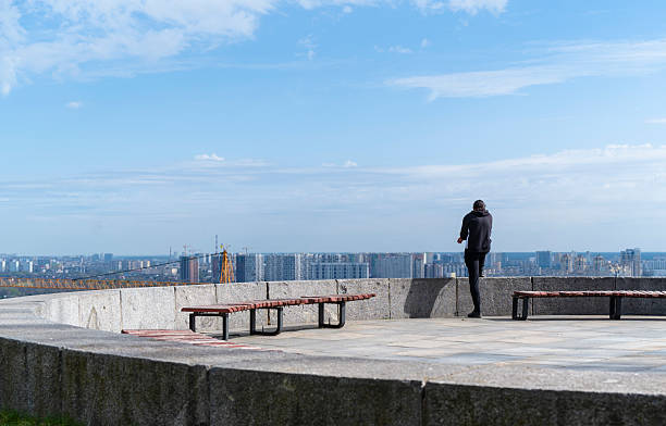 Kyiv, Ukraine - 25th April, 2025: Young adult man standing at a viewpoint, gazing at Kyiv’s cityscape with cranes and residential buildings on a sunny spring day