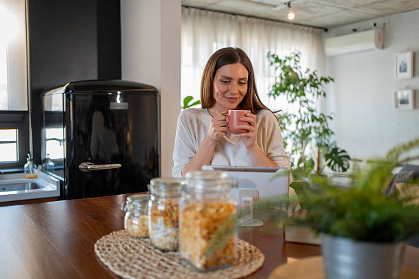 istock Smiling woman drinking coffee and using digital tablet in kitchen 2214585165