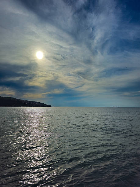 The photo shows a sunset over the sea with a clear blue sky and white clouds. Sunlight gently pours on the water surface, and in the distance you can see part of the city on the mountain.
