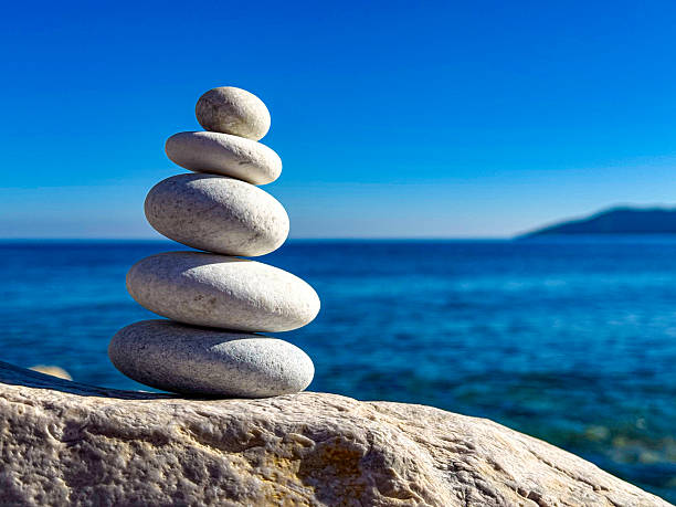 CLOSE UP: Stack of five smooth white pebbles is perfectly balanced on a stone surface by the sea. Calm blue ocean wavy and clear sky create a tranquil summer backdrop, evoking peace and mindfulness.
