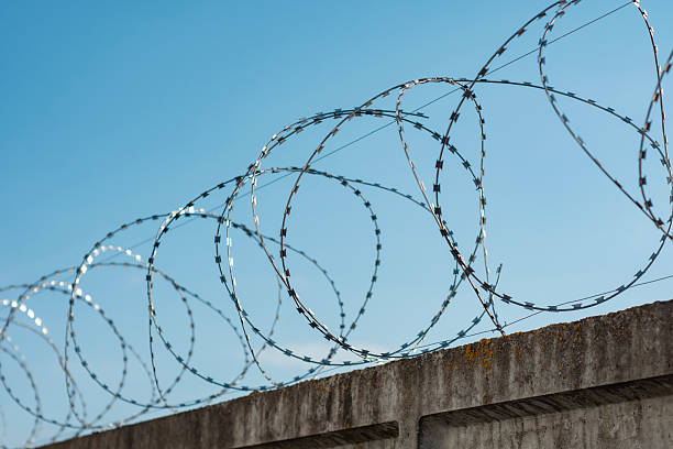 istock Coiled barbed wire fence sits atop a concrete wall under a clear blue sky, emphasizing security and restriction. 2214593536