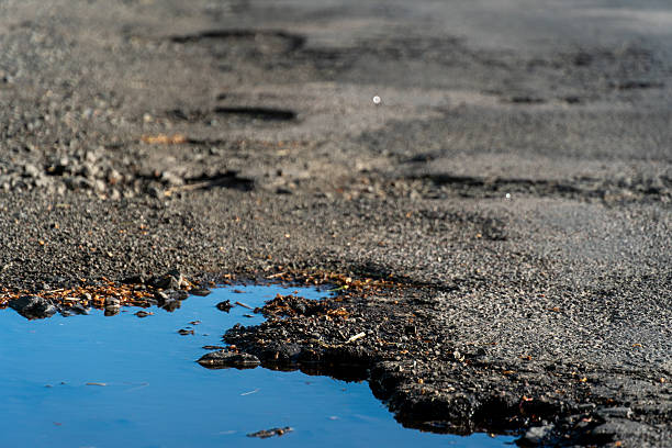 istock Standing water puddle on worn asphalt surface, rocks and debris visible along water's edge, close up 2214595772