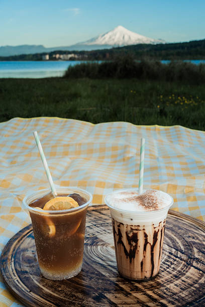 Two refreshing drinks, one with lemon and ice and the other with cream and chocolate, sit on a wooden tray on a picnic blanket with scenic lake and volcano view