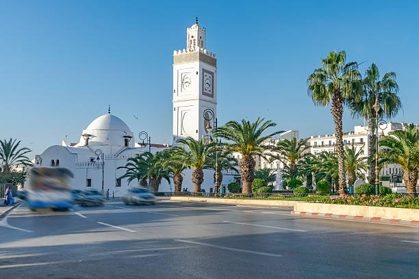 The old great mosque, El Hadi Mghiref garden, blurred traffic and people, sunlit clock minaret and buildings, palm trees, and a blue sky on a sunny day.