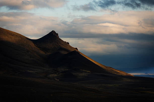A dramatic volcanic peak in the Icelandic highlands is illuminated by the last rays of golden hour light, casting striking shadows across the rugged terrain under a moody sky.