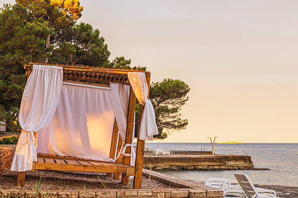 A wooden cabana draped in soft white fabric invites relaxation by serene waters as the sun sets. The tranquil beach setting offers a perfect escape for peaceful moments in nature.