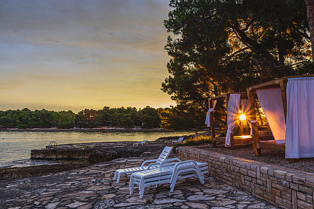 At a picturesque seaside retreat, lounge chairs rest on sun-kissed stone as the warm sun sets behind trees. Soft breezes carry hints of relaxation and tranquility along the shoreline.