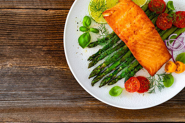 istock Fried salmon steak with cooked green asparagus, cherry tomatoes and lemon slices served on white plate on wooden table 2214621836