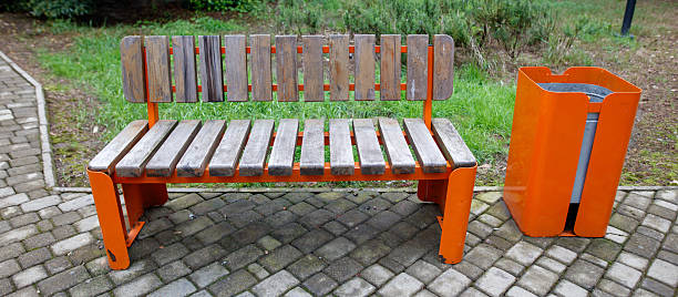 A bench and trash can sit in a park. The bench is orange and made of wood. The scene is peaceful and quiet, with no people visible