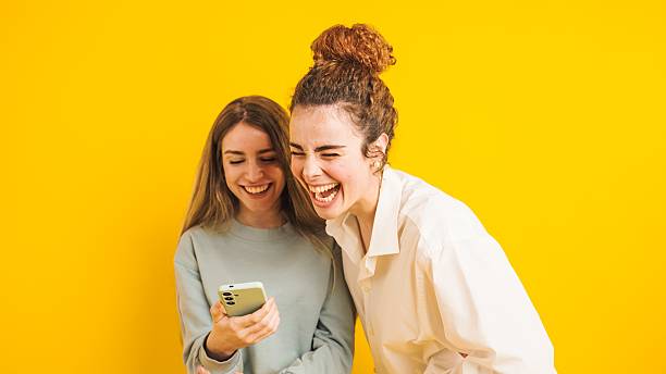 istock Two young women laughing while looking at smartphone on yellow background 2214636114