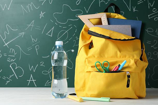 istock Backpack with different school stationery and bottle of water on white table against green chalkboard 2214636212