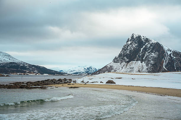 Winter coastal view at Storsandnes Beach in Lofoten, Norway, showing snowy shoreline, calm waves, and a towering rugged peak.