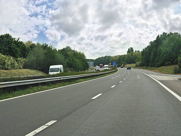 A modern multi-lane highway winds through lush green forest areas with sparse traffic visible in the distance. The asphalt road with clear white markings stretches ahead under a dramatic sky filled with fluffy white clouds, creating an inviting journey through the verdant countryside.