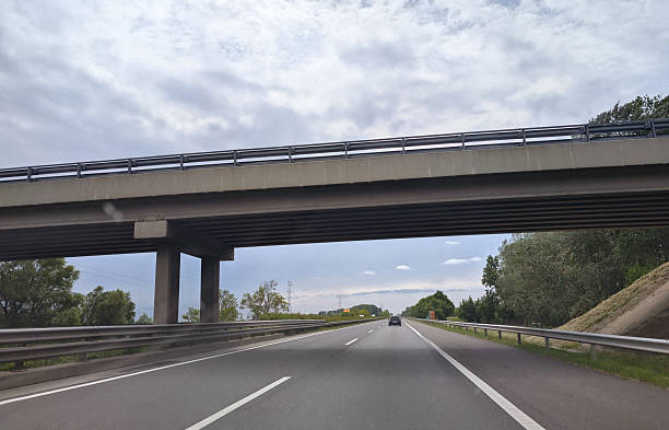 A multi-lane highway stretches into the distance beneath a large concrete overpass. The road is nearly empty with only one visible car ahead, while dramatic cloudy skies create a moody atmosphere above the transportation infrastructure.