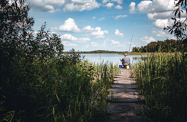 A bright yellow fishing rod extends into the frame, its tip and line in crisp focus against the vibrant, blurred backdrop of a shimmering alpine lake. The turquoise water glows with patches of green and blue, bordered by lush grasses and smooth shoreline rocks. Towering evergreen trees line the distant shore under a brilliant blue sky, creating a tranquil scene perfect for anglers and nature lovers alike.