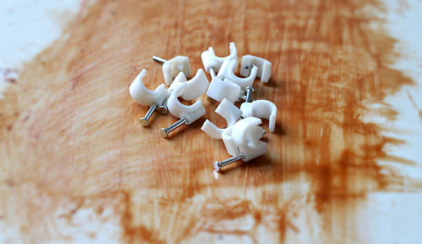 Rough slightly dusty background White color plastic or PVC material U shape clamp or cable clip, used to hold wires or electric tubes or pipes with metal nail over rough slightly dusty acrylic table background with  brown wood varnish or epoxy resin paint. Apt for construction or DIY home improvement hardware posters, banners.