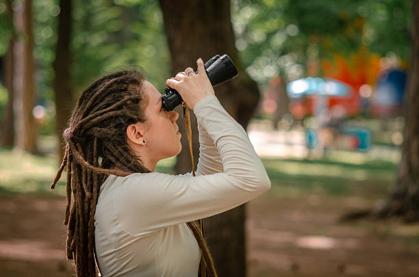 A nature-loving woman observes birds through binoculars, spotting nests hidden among leafy branches. The setting is peaceful, perfect for outdoor exploration and birdwatching hobbies.