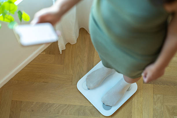 Female standing on a digital smart scale while using her smartphone to monitor weight and fitness progress at home.