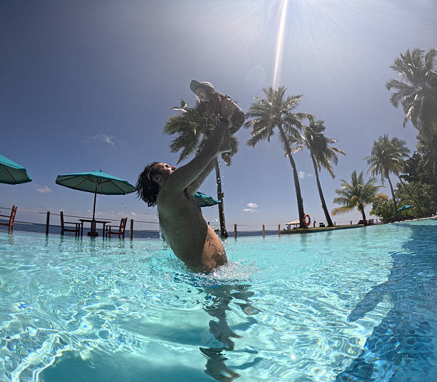 Dad playfully lifting his baby high above the stunning turquoise water of a Maldives infinity pool, surrounded by palm trees