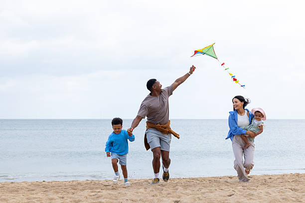 Wide shot of a family of four having fun at Seaton Sluice beach in Northumberland, North East England. The mother is holding a baby girl on one hip, while the father holds hands with a young boy, flying a colourful kite in the other hand. They are all running along the sand, wearing casual summer clothing and there is an expanse of calm sea behind them.\n\nVideos similar to this scenario available.